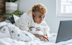 focused black woman typing on laptop while resting on bed